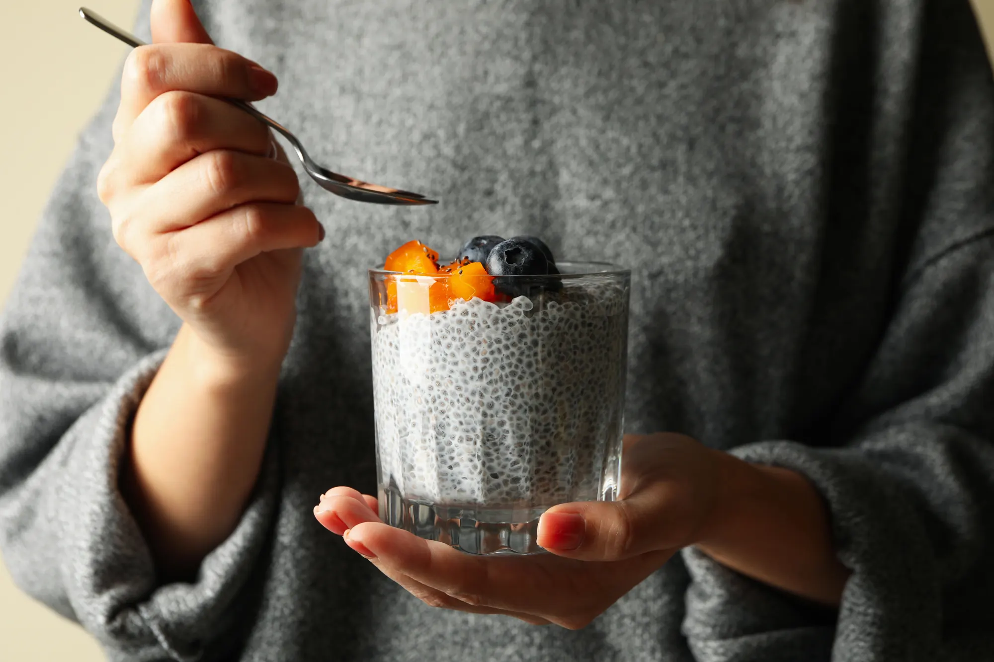 Une femme en pull gris mange un pudding de chia aux fruits dans un verre